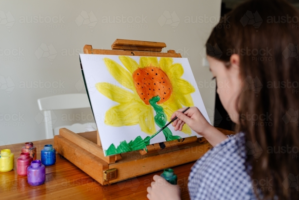 Young girl at home sitting at an easel painting a sunflower - Australian Stock Image