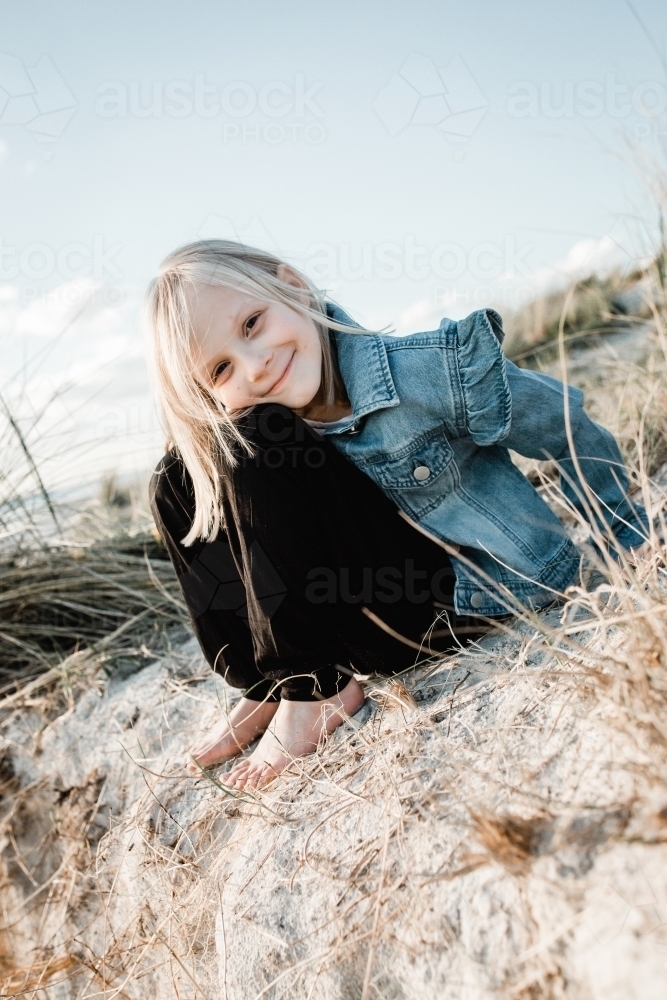 Young girl at the beach sitting on a sand dune looking at the camera wearing a denim jacket - Australian Stock Image