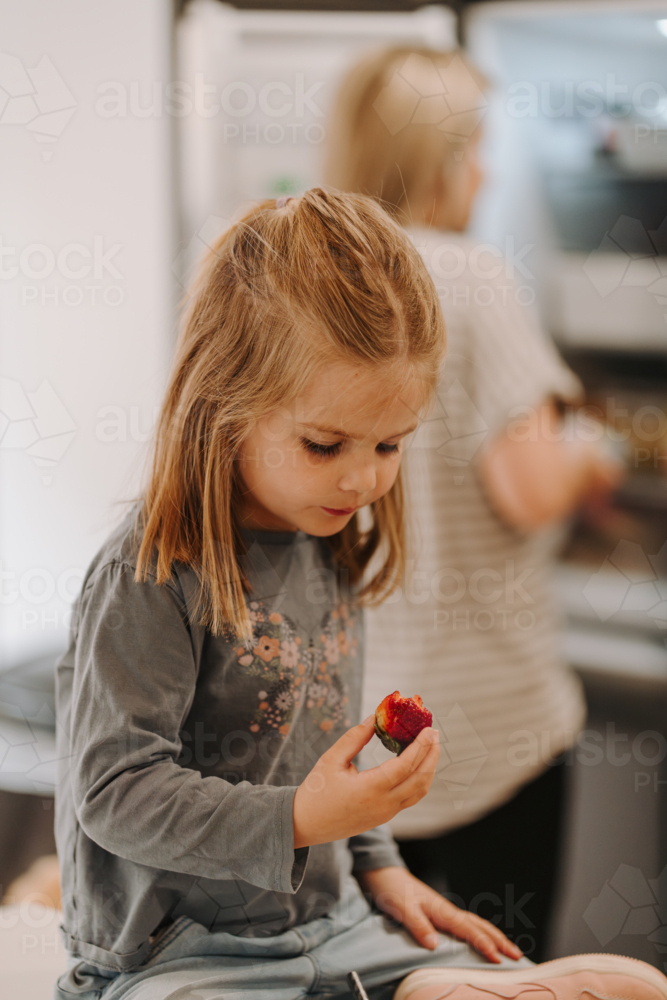 Young girl eating strawberry while sitting on the countertop. - Australian Stock Image