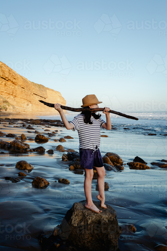 Young girl having fun playing at the beach standing on a rock holding a branch like a spear - Australian Stock Image