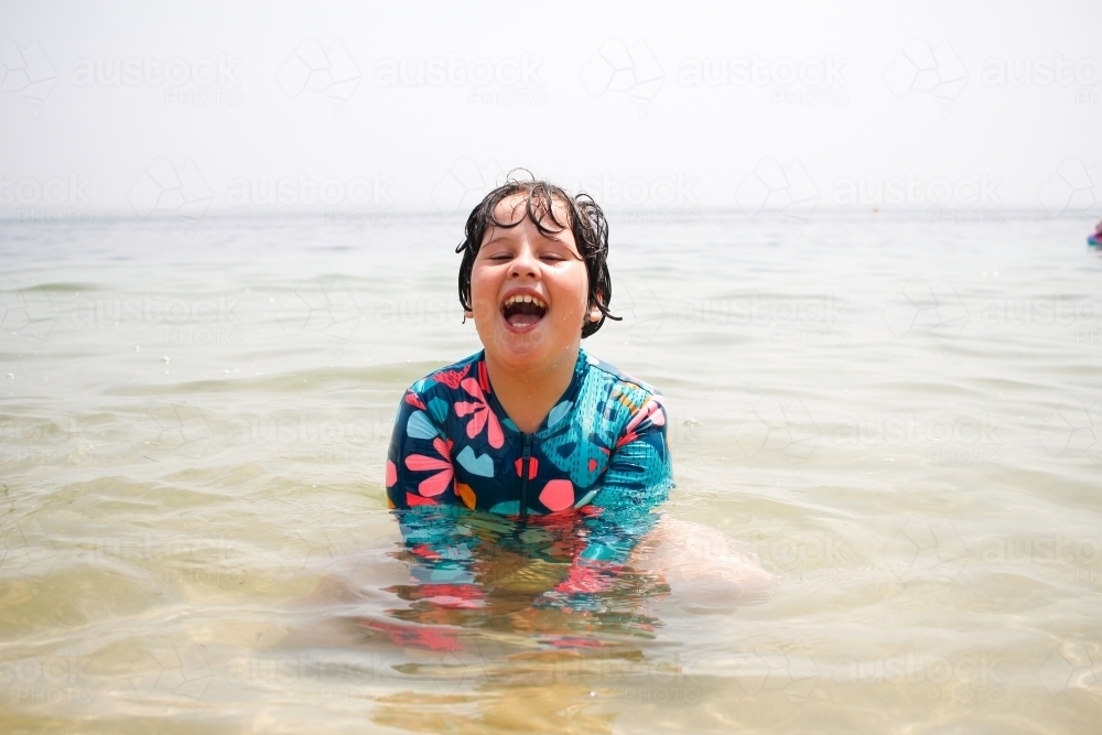 Young girl laughing as she sits in the calm shallows at the beach - Australian Stock Image