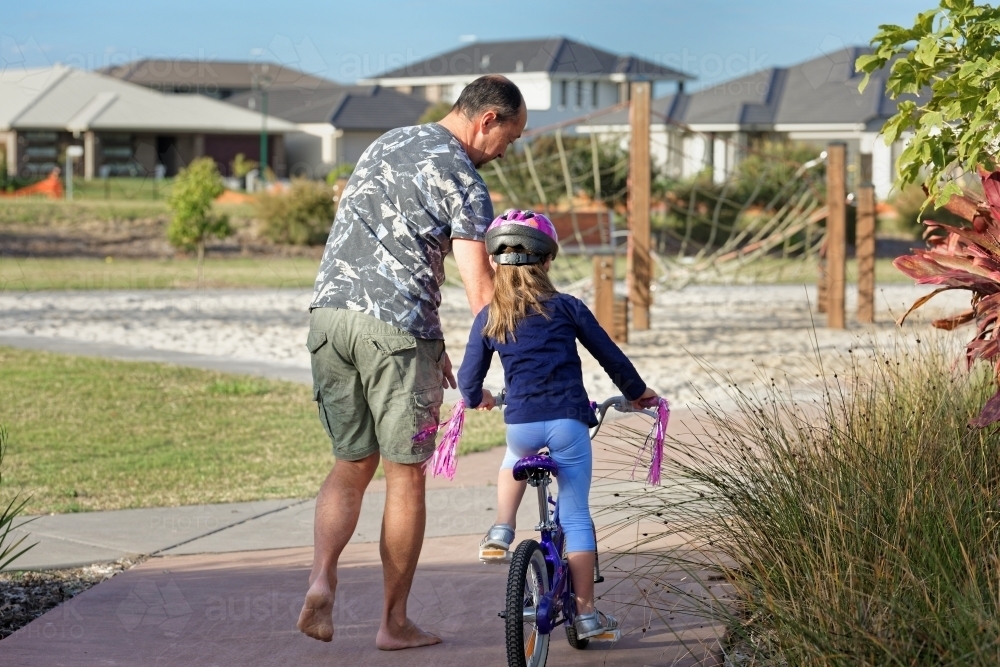 Young girl learning to ride her bicycle in the park with her dad helping - Australian Stock Image