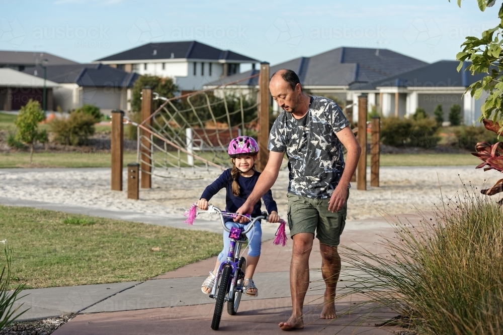 Young girl learning to ride her bike with her dad in the park without training wheels - Australian Stock Image