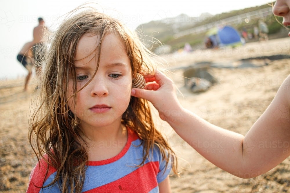 Young girl listening intently as older girl holds a shell to her ear at the beach - Australian Stock Image