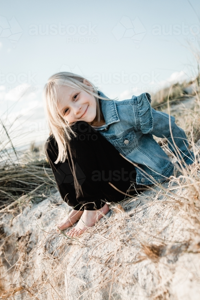 Young girl wearing a denim jacket at the beach sitting on a sand dune - Australian Stock Image