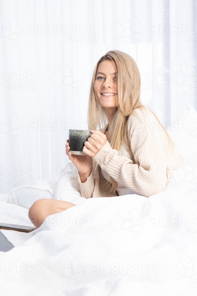 Young lady drinking hot drink in white bed - Australian Stock Image