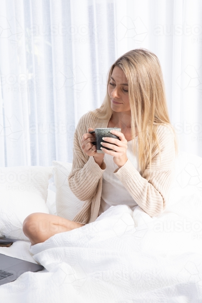 Young lady drinking hot drink in white bed - Australian Stock Image