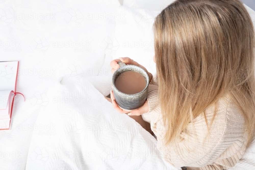 Young lady drinking hot drink in white bed from an overhead perspective - Australian Stock Image