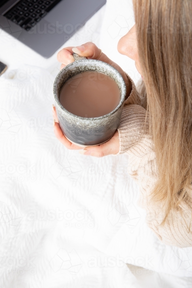 Young lady drinking hot drink in white bed from an overhead perspective - Australian Stock Image