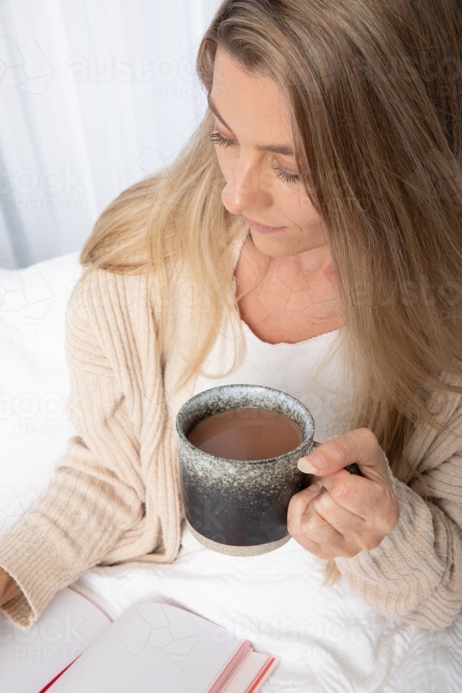 Young lady drinking hot drink in white bed writing in a journal - Australian Stock Image