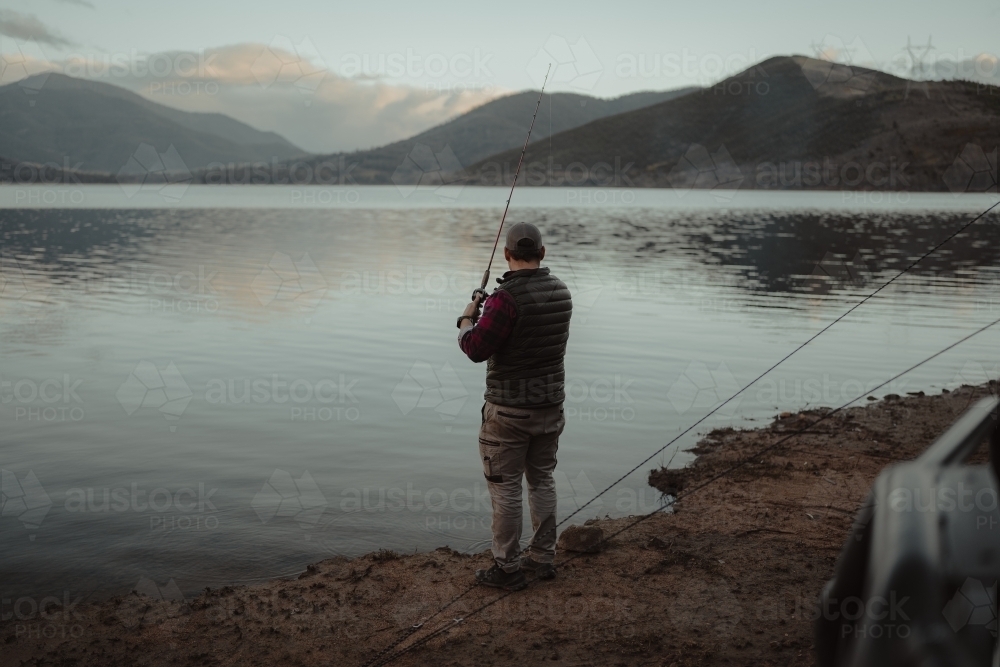 Young man casting a fishing line into a calm lake with mountains in the background - Australian Stock Image