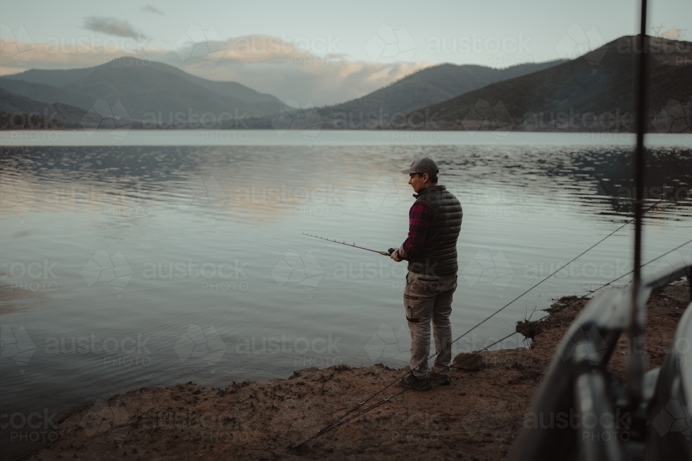 Young man fishing next to a calm lake with mountains in the background - Australian Stock Image