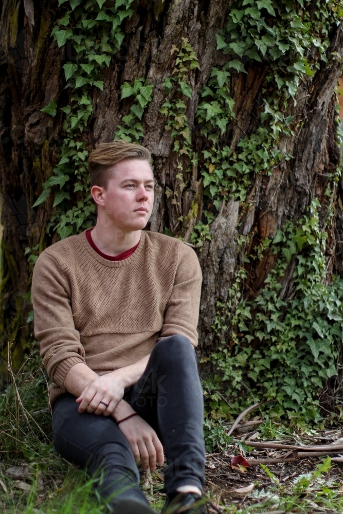 Young man sitting outdoors at the base of a tree contemplating life - Australian Stock Image