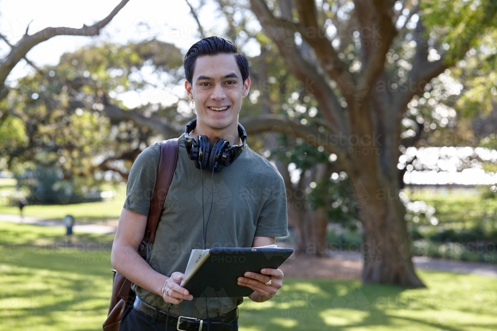 Young man smiling outside holding a device with headphones at park - Australian Stock Image