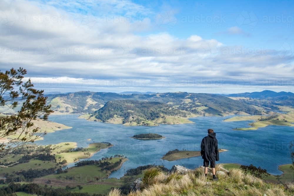 Young man standing on a hill overlooking blue lake - Australian Stock Image