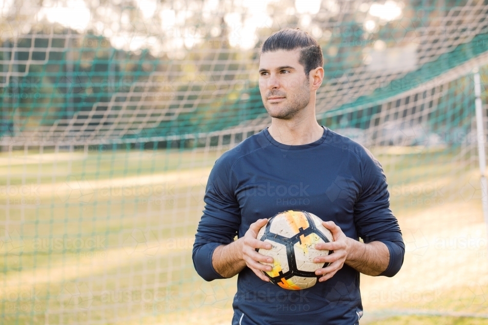 Young man standing on the field while looking at his left side, holding a soccer ball near the net - Australian Stock Image