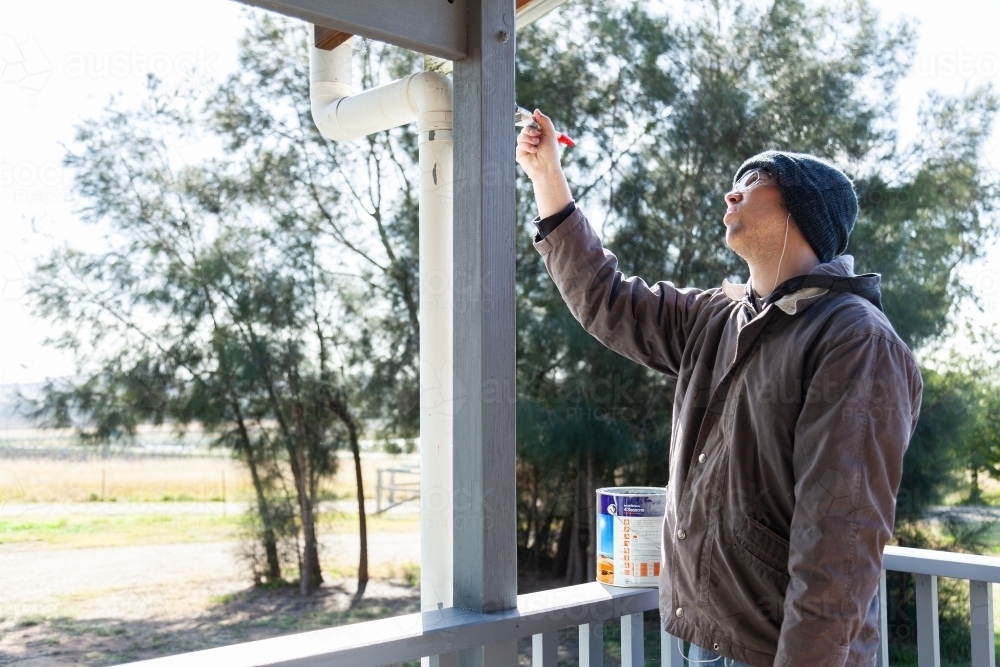 Young man tradie painting railing on house in morning with grey paint - Australian Stock Image