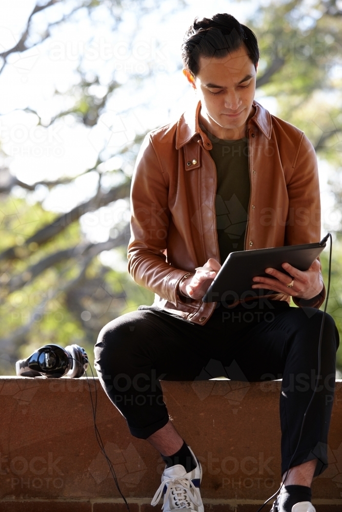 Young man with dark hair outside holding device with headphones at park - Australian Stock Image