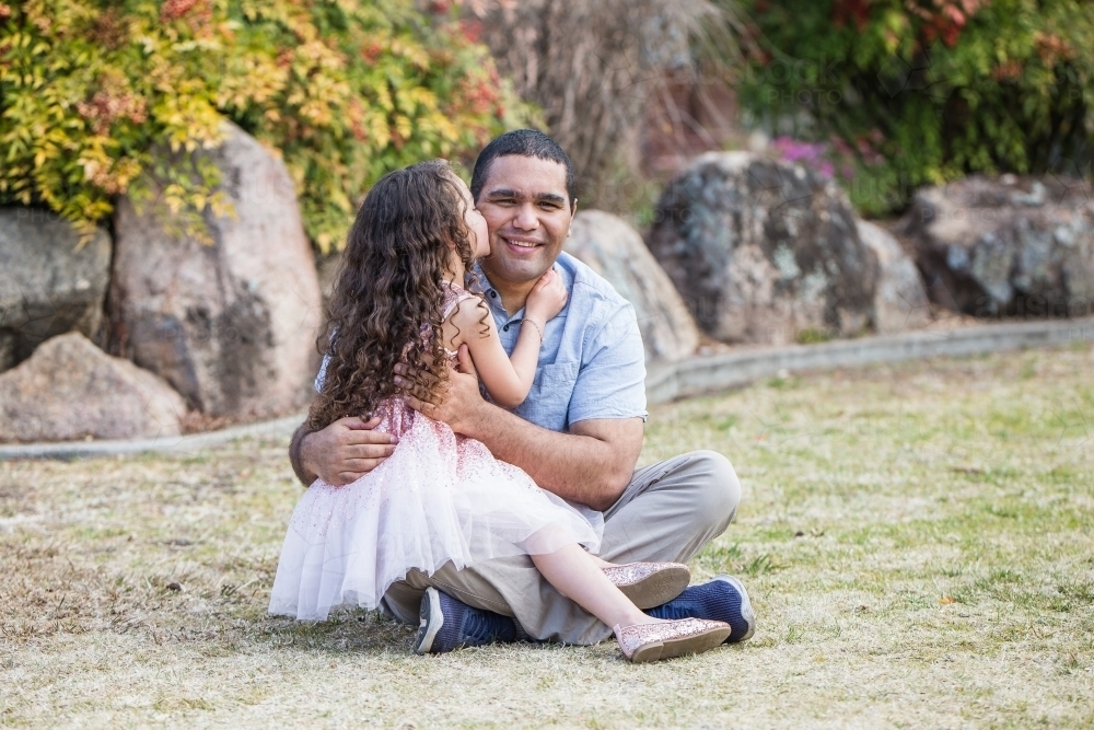 Young mixed race aboriginal caucasian girl sitting on father's lap kissing cheek - Australian Stock Image