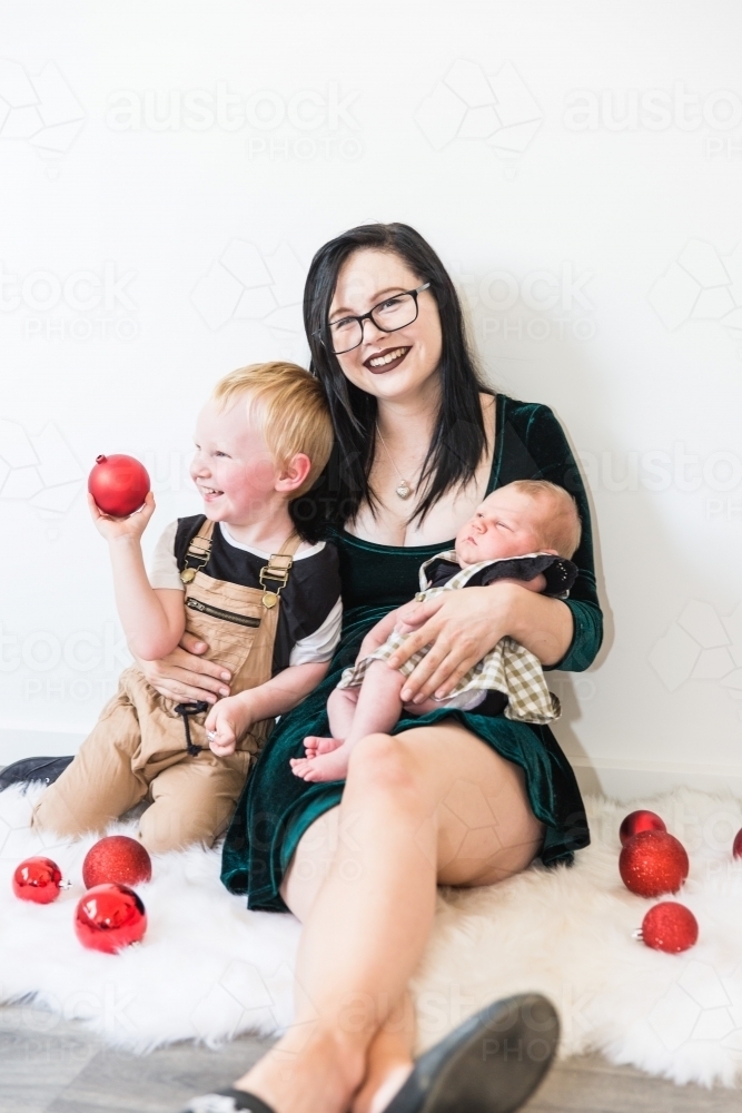 Young mother sitting on rug holding her two children at Christmas with decorations - Australian Stock Image