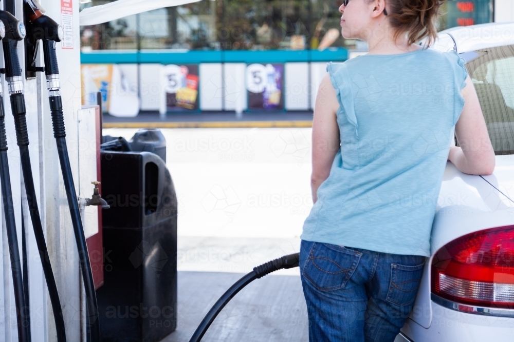 Young person filling up car with fuel at petrol station leaning on car - Australian Stock Image