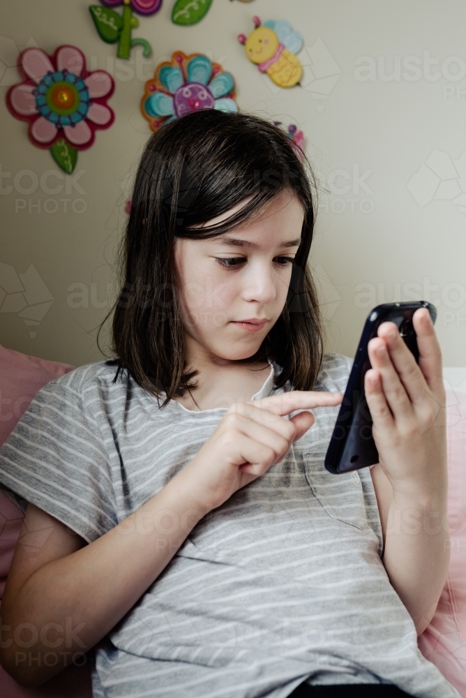 Young preteen girl in her bedroom sitting on bed texting chat messages on mobile phone / smartphone - Australian Stock Image