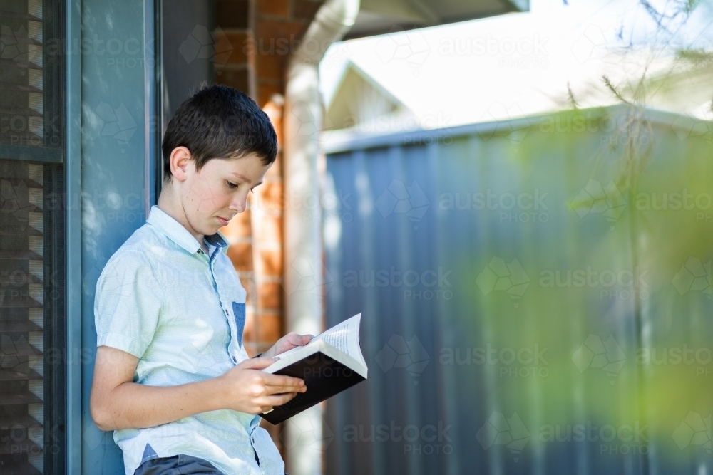 Young primary aged boy reading fiction book leaning against window outside home - Australian Stock Image