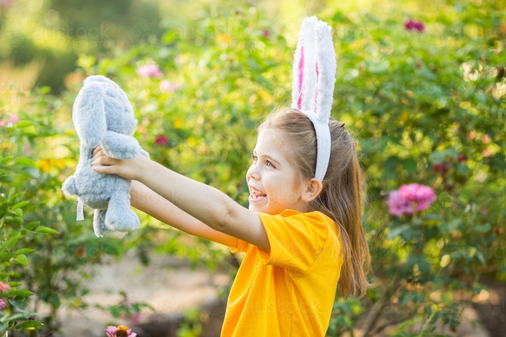 Young school girl standing outside in garden at Easter time holding out her bunny rabbit toy - Australian Stock Image