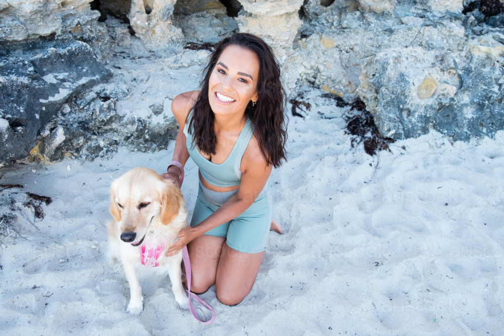 Young smiling woman in active wear kneeling next to dog on beach - Australian Stock Image