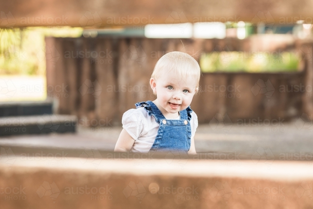 Young toddler girl peeking through gap in garden wall - Australian Stock Image