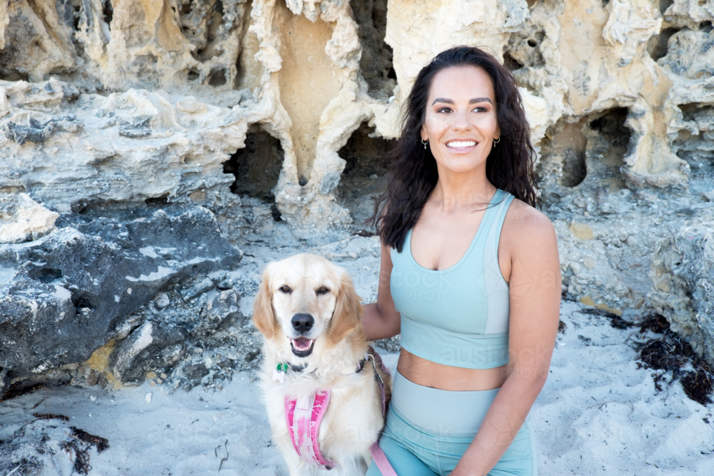 Young woman in active wear kneeling next to large golden dog on beach - Australian Stock Image