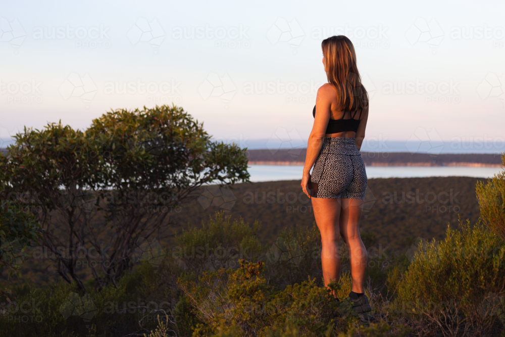 Young woman in fitness wear looking at scenic view over the sea from a mountain lookout at sunset - Australian Stock Image