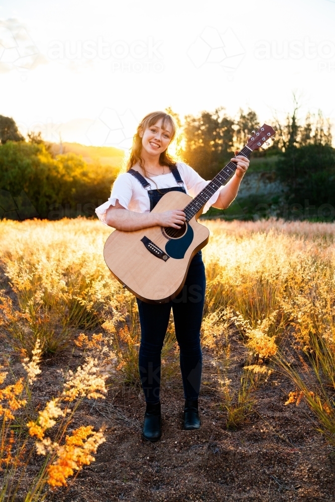 Young woman playing guitar in grass backlit by golden light - Australian Stock Image