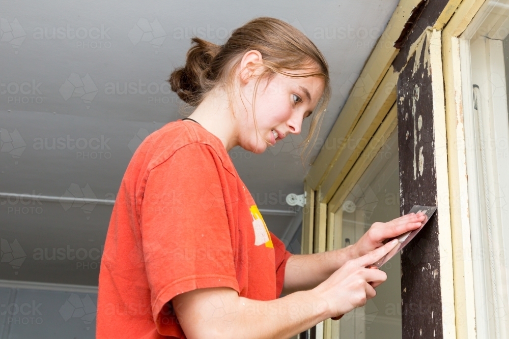 Young woman scraping paint off window frame - Australian Stock Image