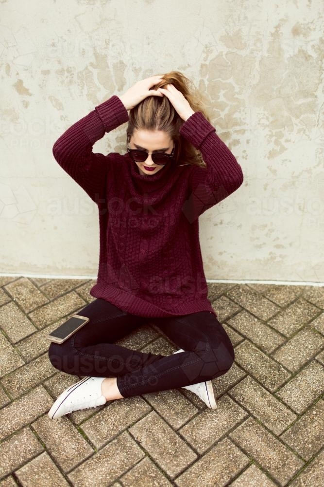 Young woman sitting against a wall with a phone and sunglasses - Australian Stock Image