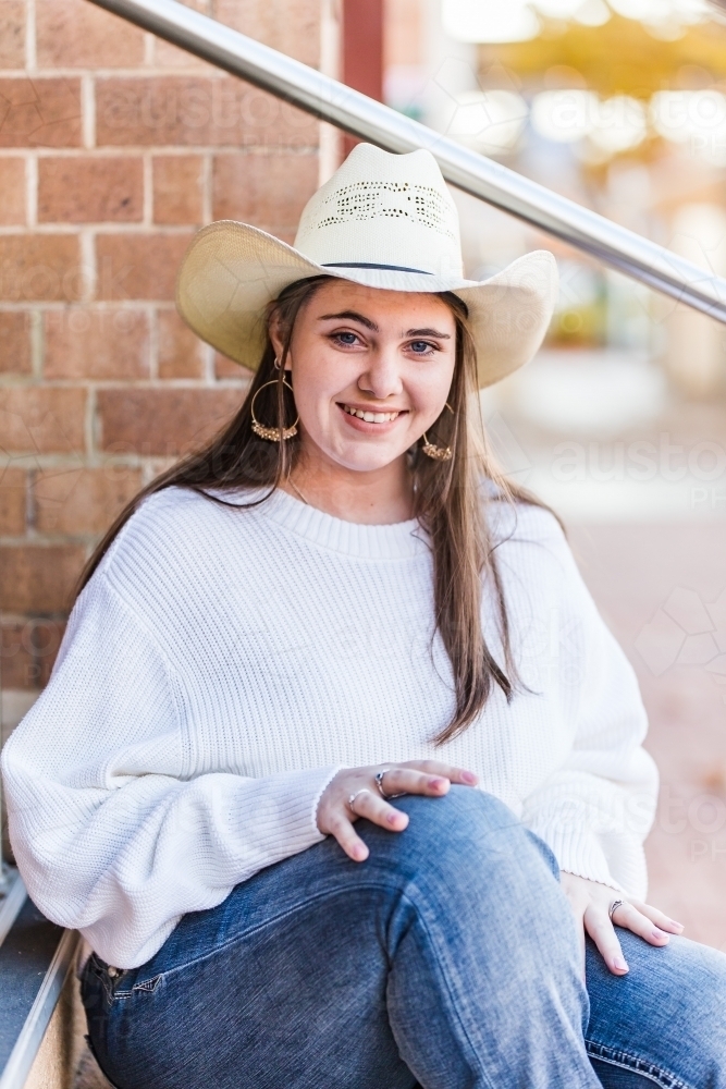 Young woman sitting with hands on knees wearing hat smiling - Australian Stock Image