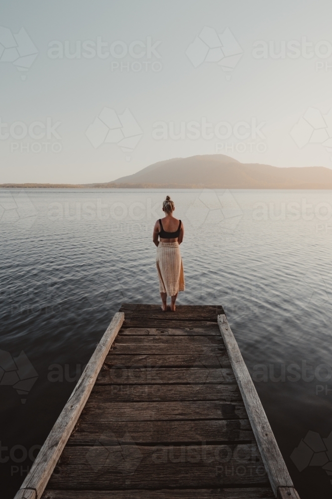 Young woman standing at the end of a jetty looking over a lake with a mountain in the background. - Australian Stock Image