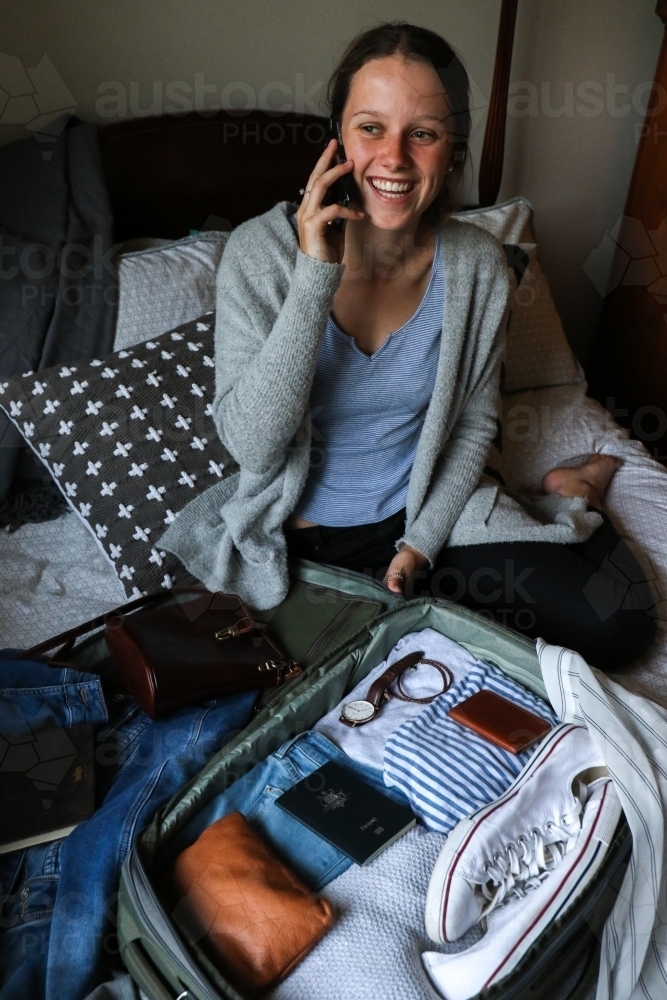 Young woman talking on the the phone next to her packed bag ready to travel - Australian Stock Image