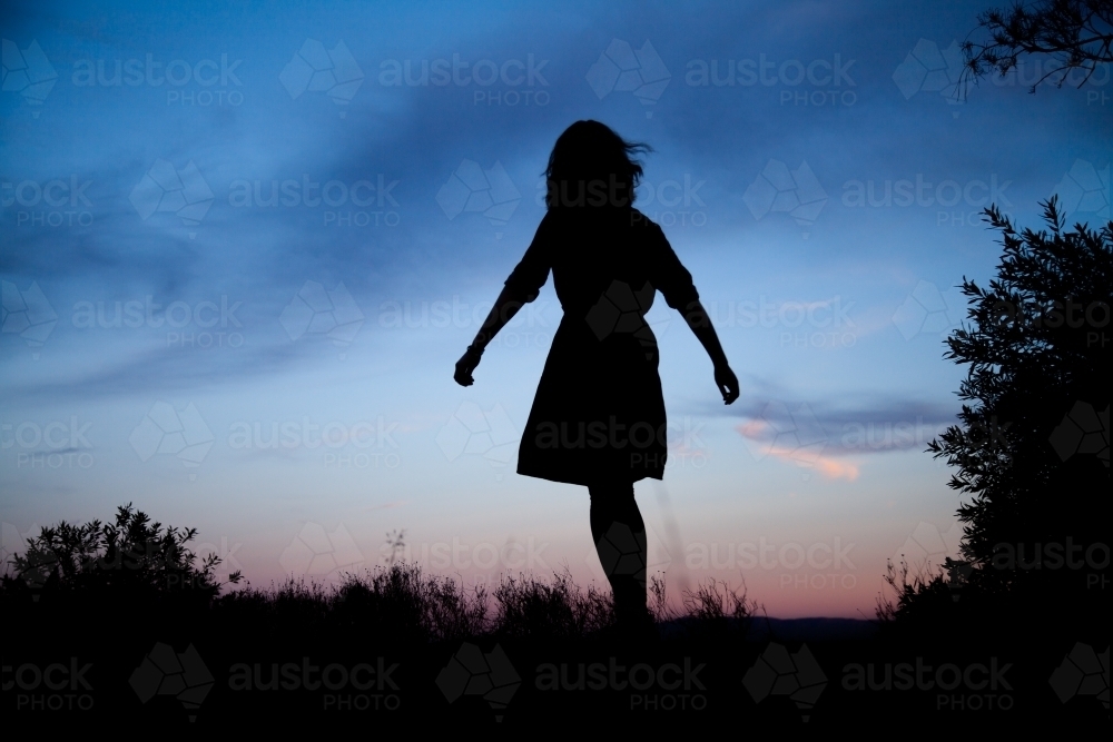 Young woman twirling silhouetted against pastel dusk sky - Australian Stock Image