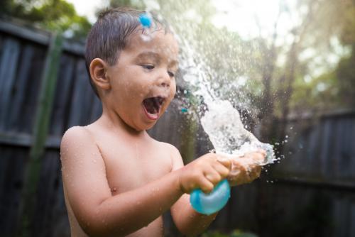 3 year old mixed race boy plays excitedly with water bombs in suburban backyard - Australian Stock Image