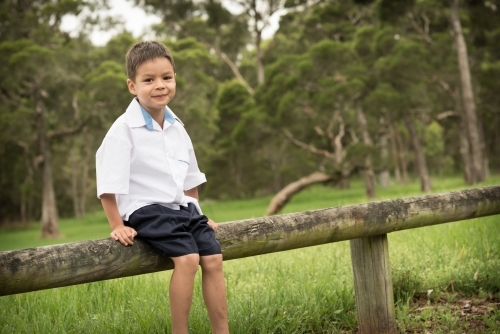 6 year old mixed race boy waits on a road side fence post for his first day of school - Australian Stock Image