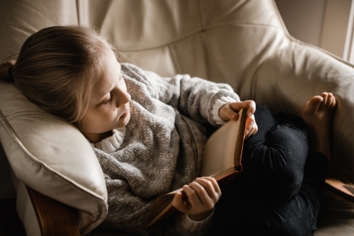A blonde caucasian girl sitting curled up on a chair reading a book - Australian Stock Image