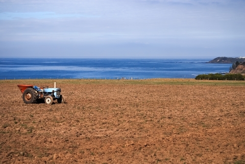 A blue and red tractor on barren farmland overlooking the ocean - Australian Stock Image