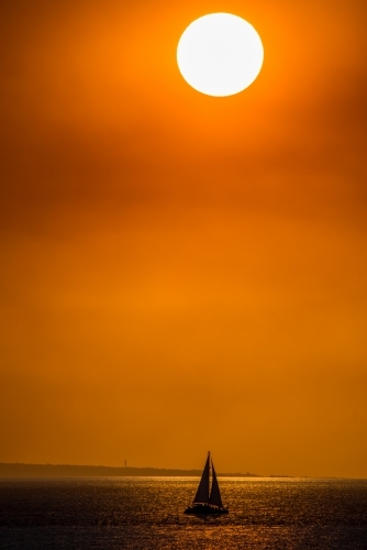 A bright sun hangs low in the sky with a small silhouette of a sailboat on the ocean. - Australian Stock Image