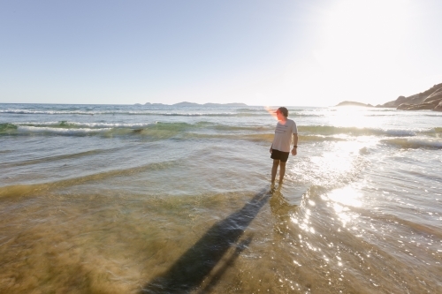 A Caucasian boy child wearing a hat standing alone in the water at an australian beach with waves - Australian Stock Image