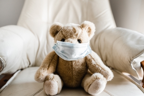 A child's teddy bear sitting on a chair wearing a facemask - Australian Stock Image