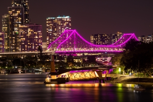 A CityCat ferry terminal on the Brisbane River with the Story Bridge lit in magenta/pink behind - Australian Stock Image