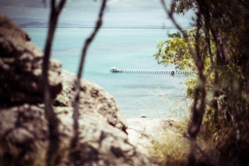 A coastal view with a long pier or jetty that can be seen through rocky formations and tree branches - Australian Stock Image