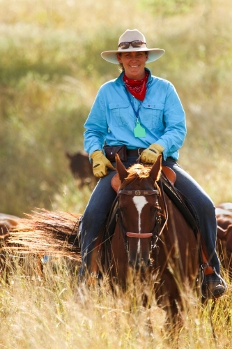 A country lady smiles at camera while mustering on horse. - Australian Stock Image