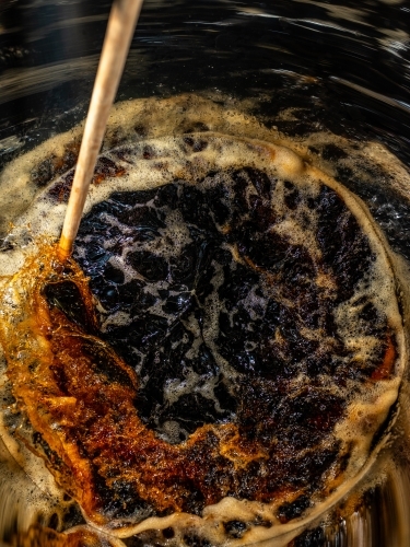 A dark liquid with frothy bubbles and swirls stirred with a wooden paddle - Australian Stock Image